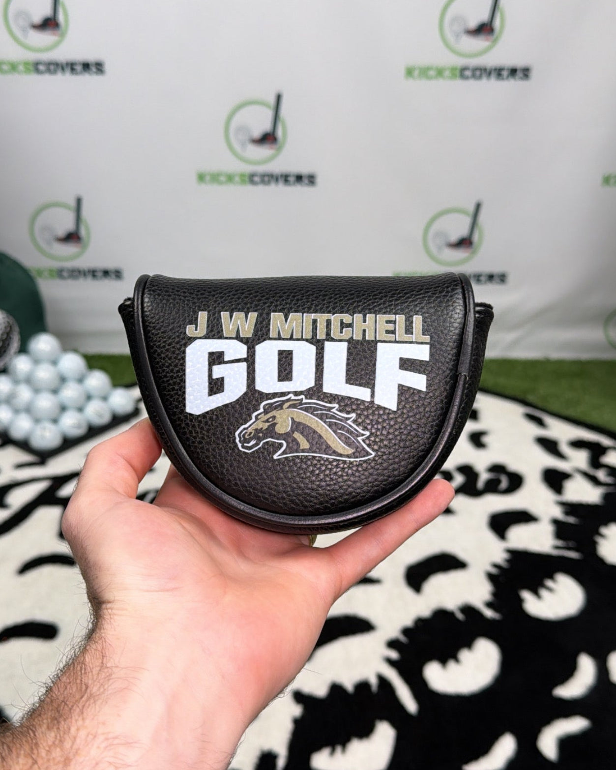 Hand holding a golf club head cover with 'J.W. Mitchell Golf' branding against a branded backdrop.