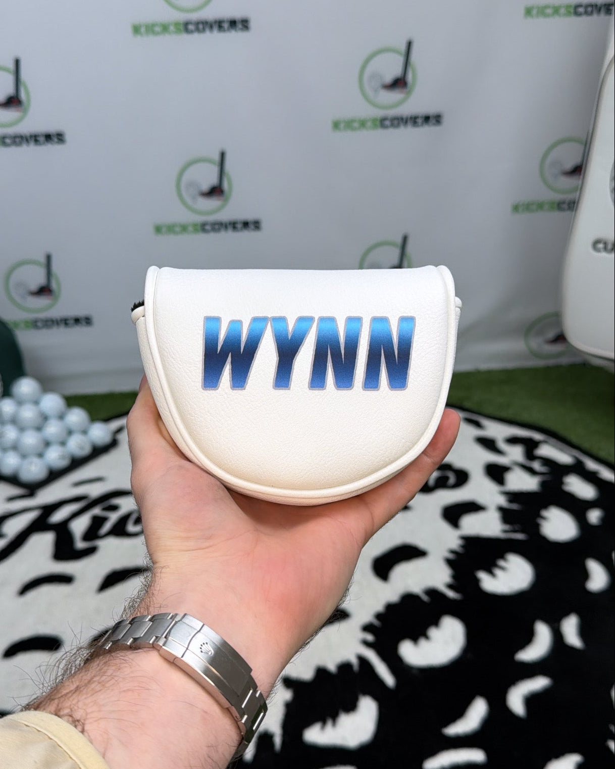 Hand holding a white golf ball marker with 'WYNN' branding against a branded backdrop.