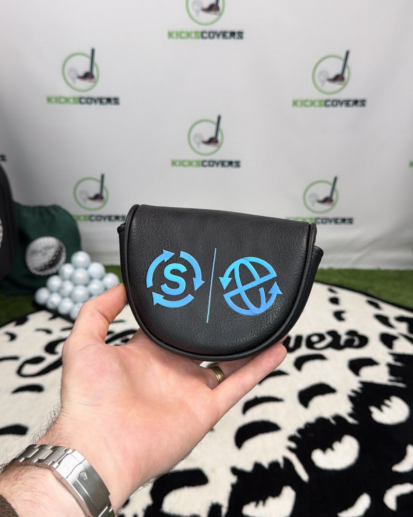 Hand holding a golf club head cover with a brand logo against a branded backdrop