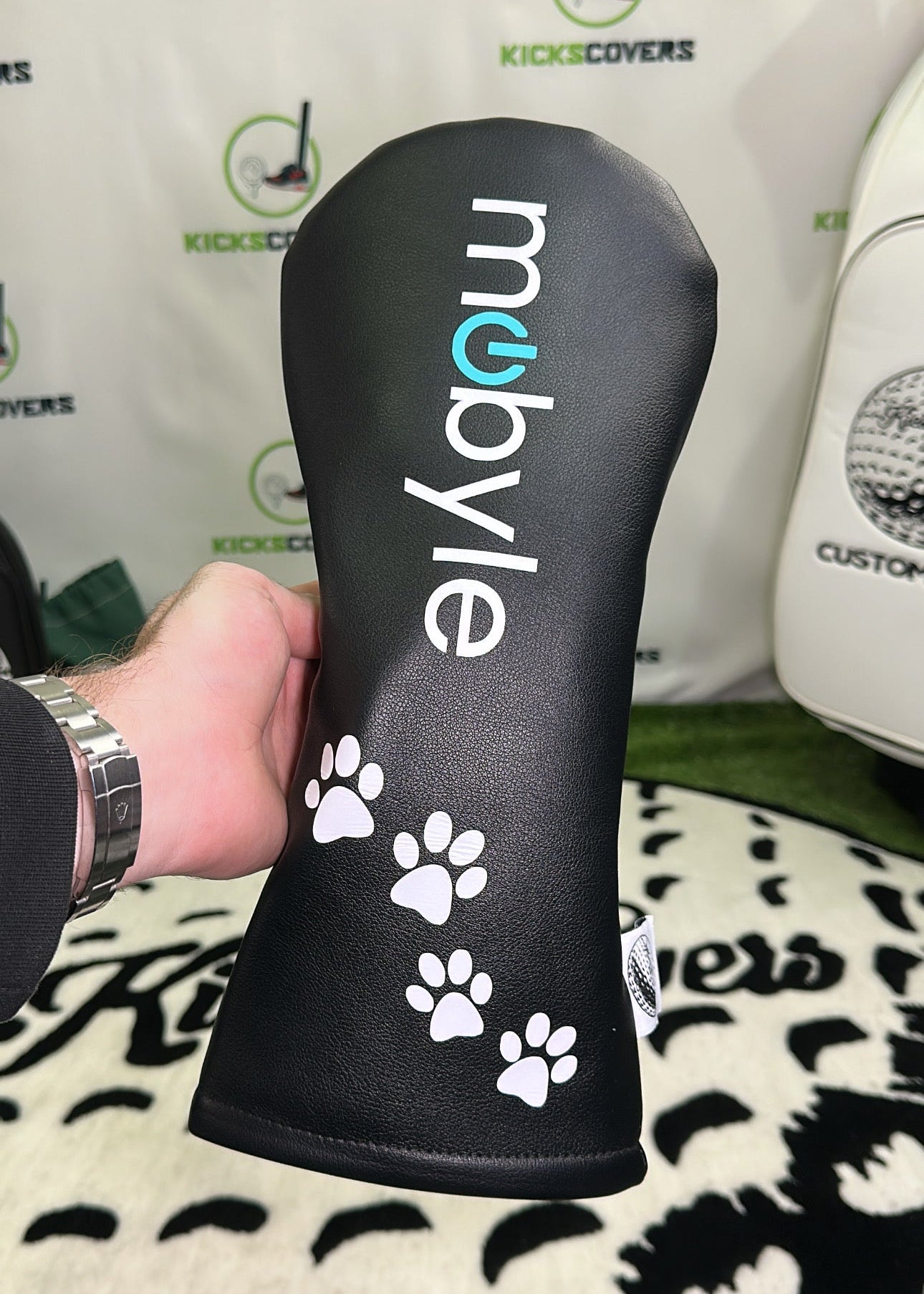 Black golf club head cover with white paw prints and 'mobyle' branding held by a person.