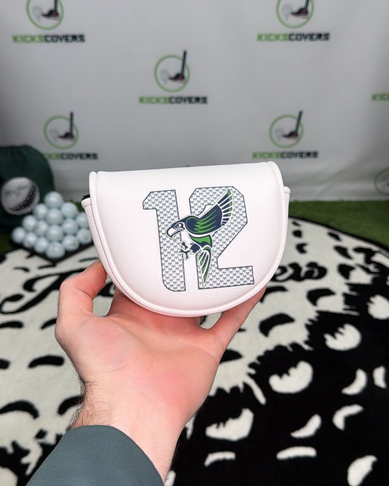 Hand holding a white golf club head cover with number 12 and logo, branded background.