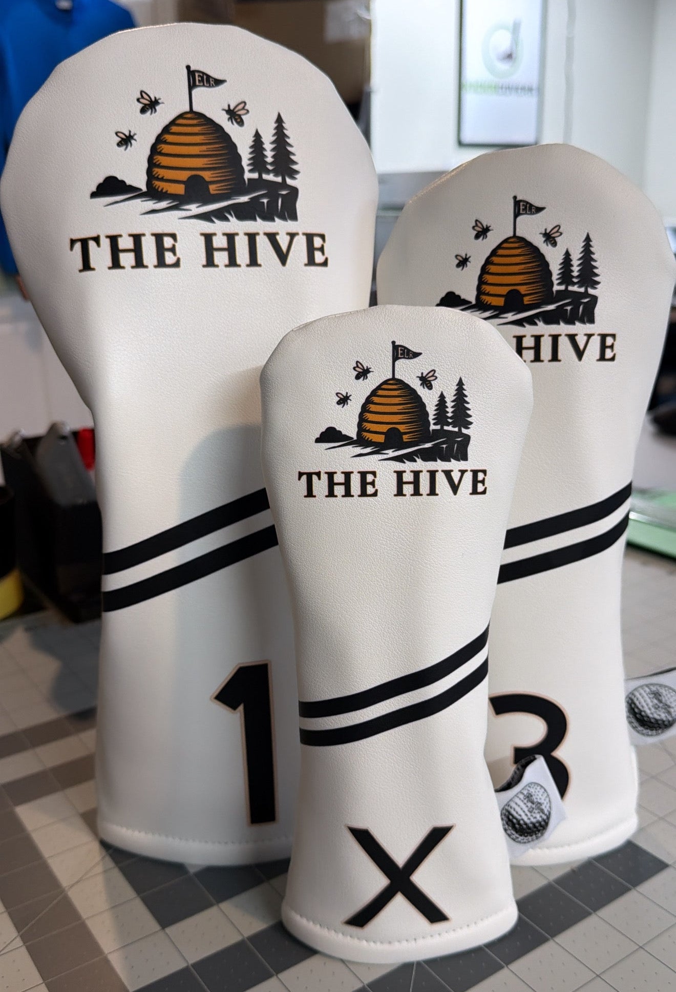 White golf club head covers with 'The Hive' logo on a tiled floor.