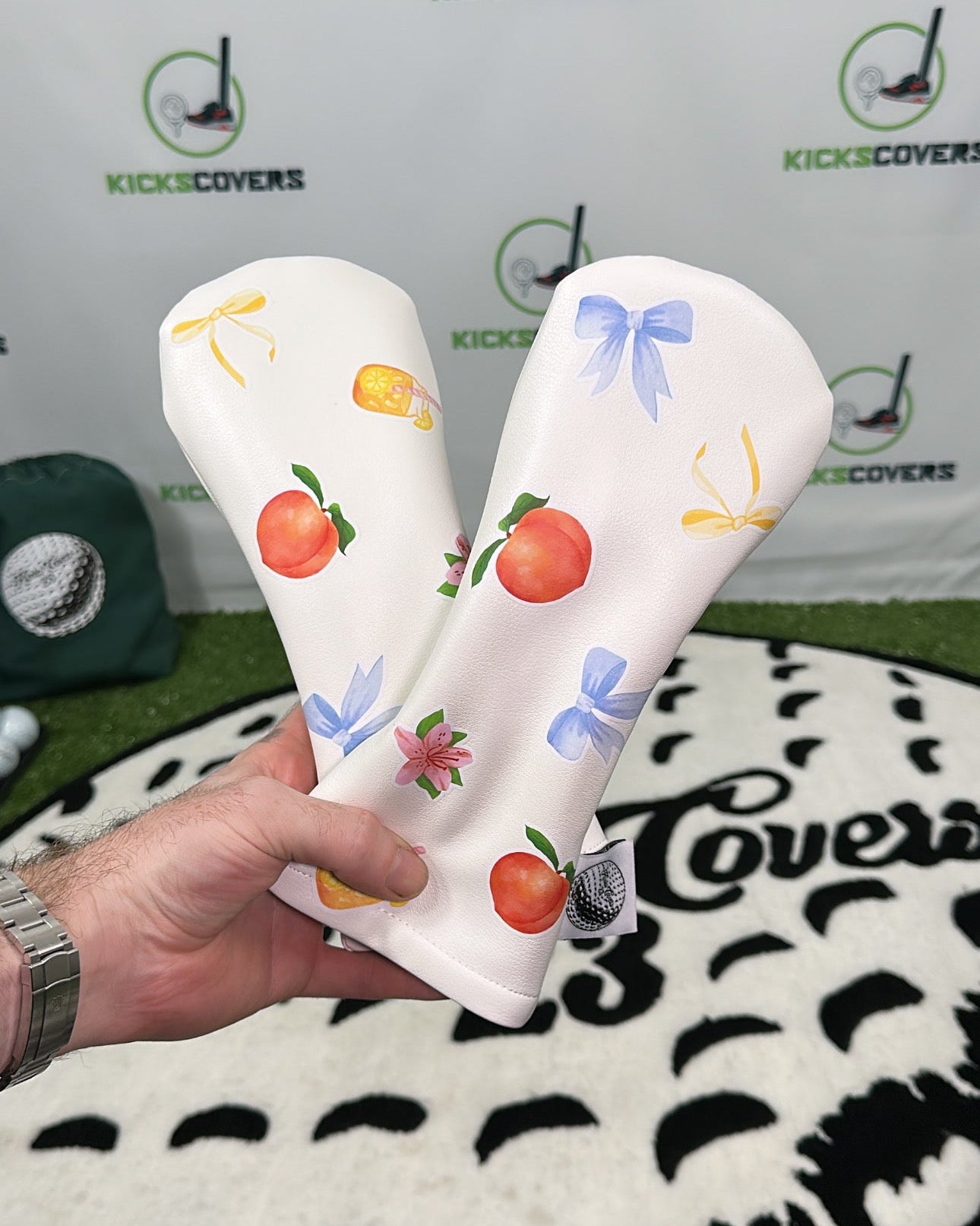 Two white headcovers with colorful designs held by a hand against a branded background.