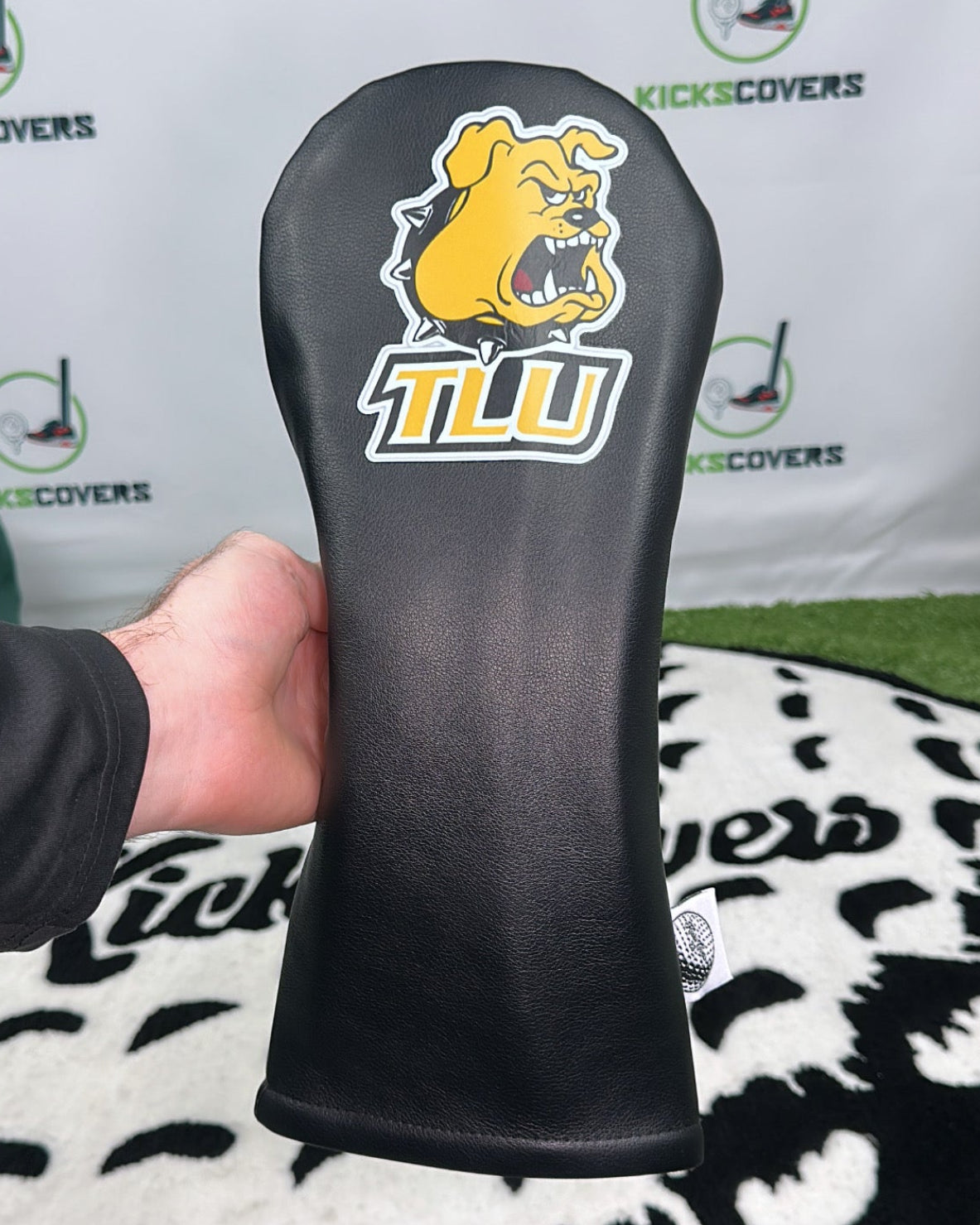 Black golf club head cover with a logo held in front of a branded backdrop