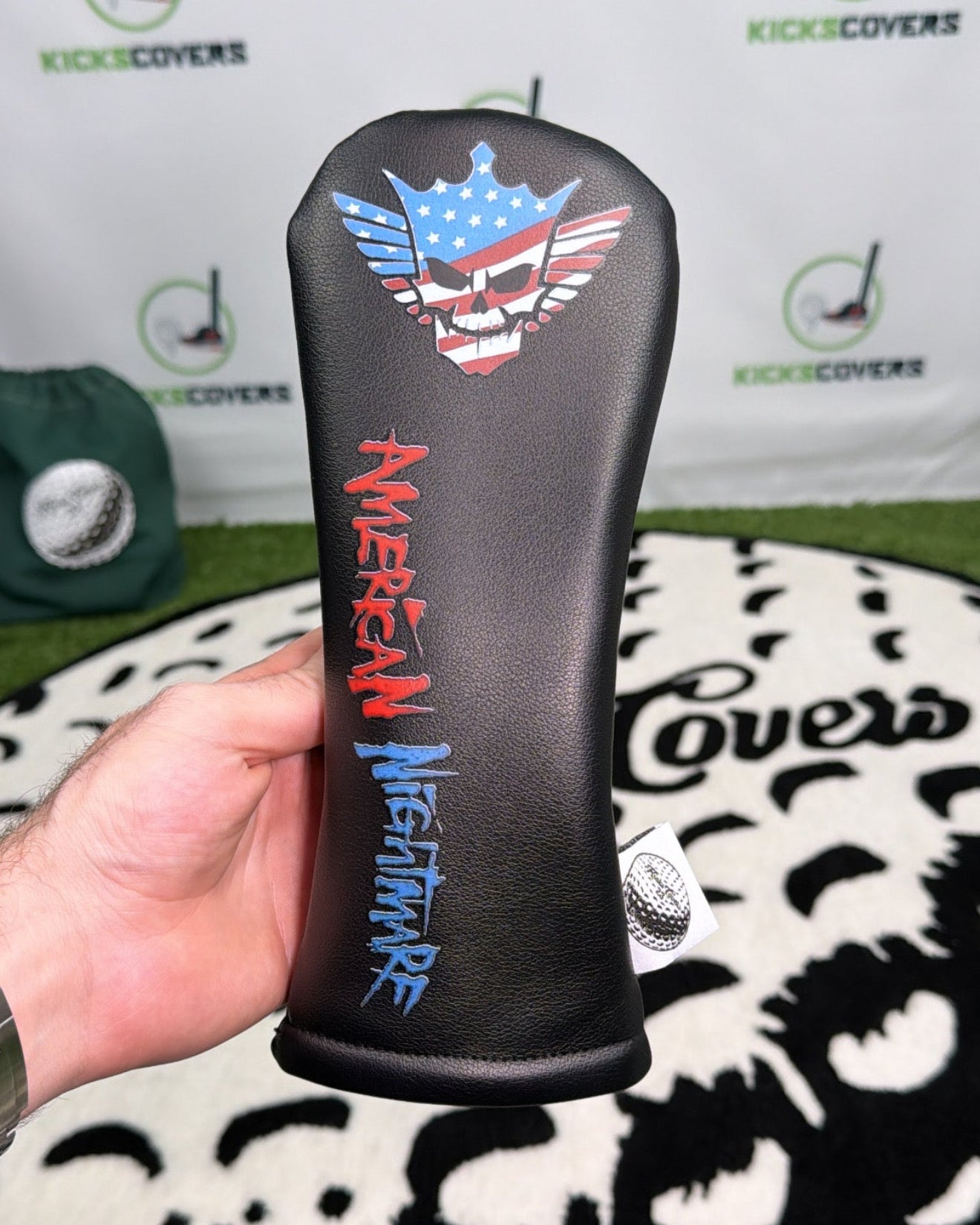 Golf club head cover with American Nightmare design held by a hand, brand 'KicksCovers' visible in the background.