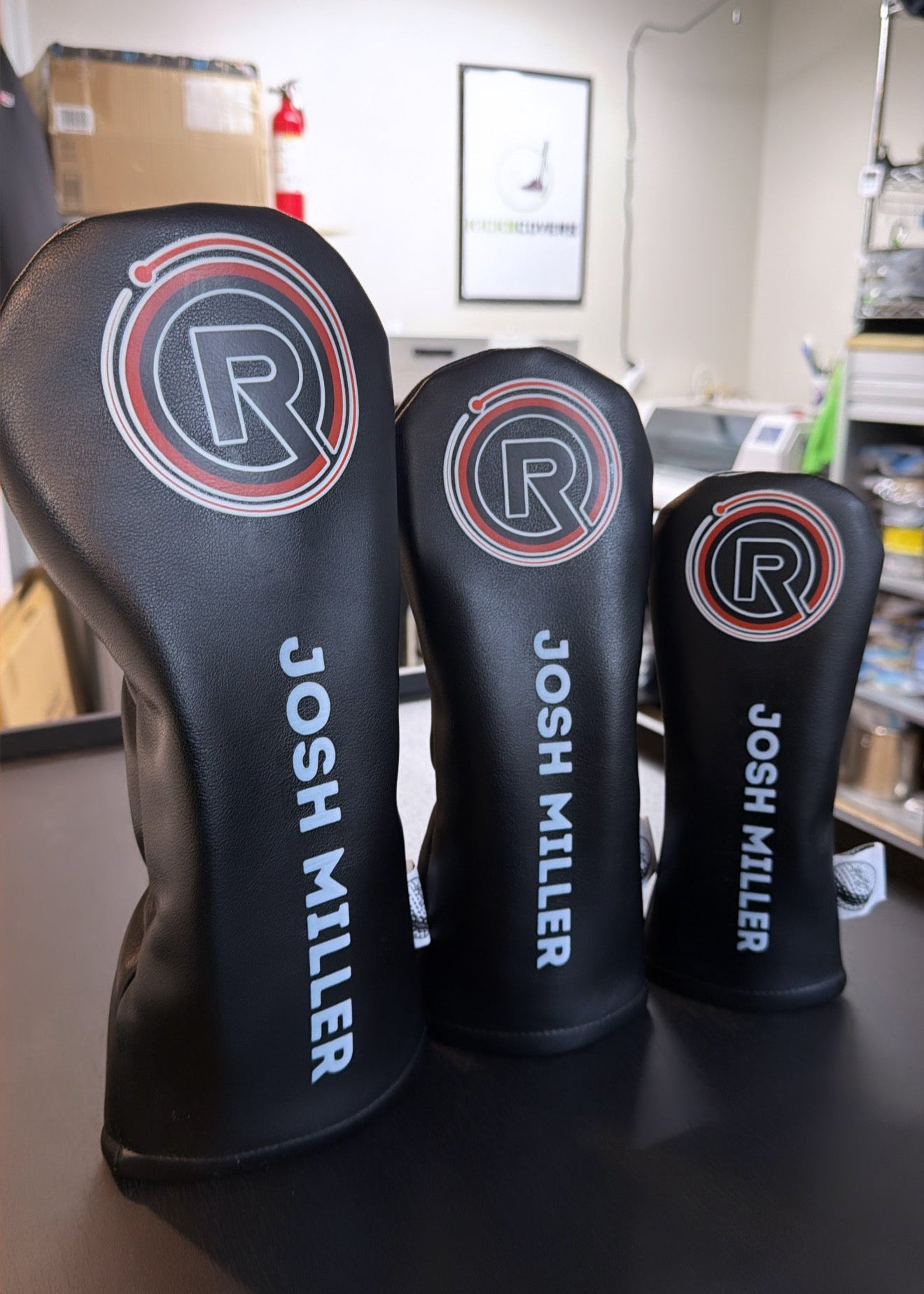 Three black golf club head covers with 'R' logo and 'Josh Miller' text on a table.